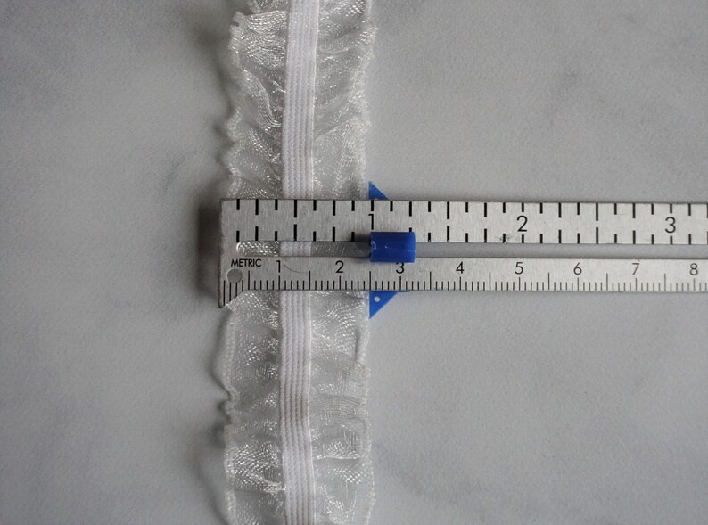 May include: Close-up of white lace trim with a ruffled edge, measured with a ruler. The trim is approximately 1 inch wide. The ruler shows both inches and centimeters for accurate measurement.