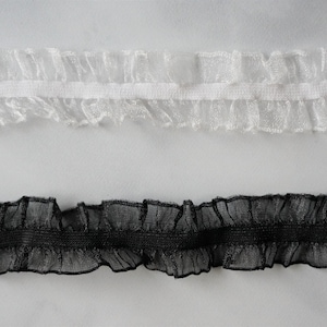 May include: Two strips of ruffled trim, one white and one black, are displayed against a white background. The trim has a delicate, sheer appearance with a ruffled edge, suitable for sewing and craft projects.