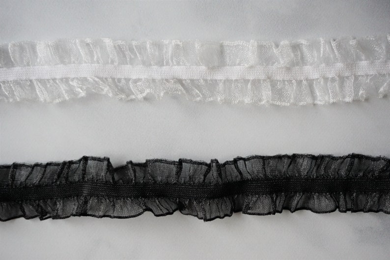May include: Two strips of ruffled trim, one white and one black, are displayed against a white background. The trim has a delicate, sheer appearance with a ruffled edge, suitable for sewing and craft projects.