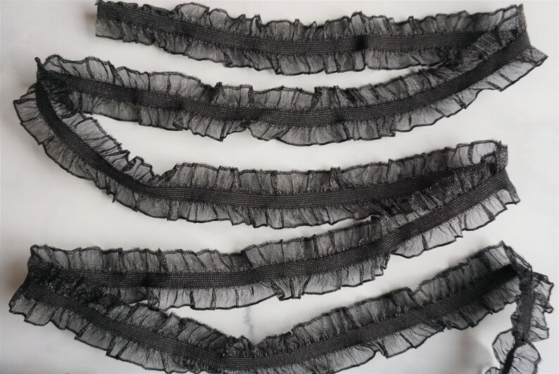 May include: Black ruffled trim ribbon. The ribbon features a sheer, ruffled edge with a darker, solid center. This decorative trim is suitable for various craft and sewing projects.