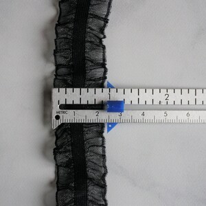 May include: Black ruffled lace trim with a central band, shown with a metric ruler. The trim is approximately 0.75 inches wide. The lace has a delicate, sheer appearance, and the ruler shows measurements in centimeters.