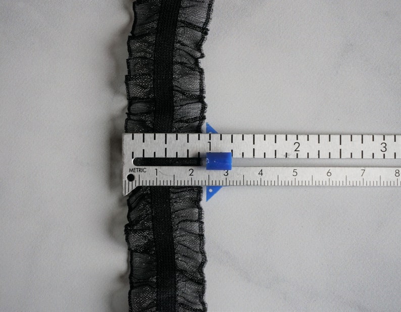 May include: Black ruffled lace trim with a central band, shown with a metric ruler. The trim is approximately 0.75 inches wide. The lace has a delicate, sheer appearance, and the ruler shows measurements in centimeters.
