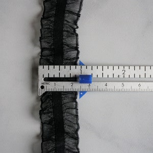 May include: Black ruffled lace trim with a central band, shown with a metric ruler. The trim is approximately 0.75 inches wide. The lace has a delicate, sheer appearance, and the ruler shows measurements in centimeters.