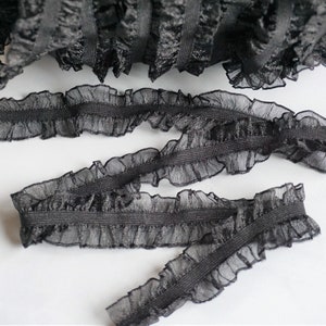 May include: Black ruffled elastic trim, perfect for sewing and crafting projects. The trim features a delicate, sheer fabric with a ruffled edge and a sturdy elastic base. The trim is shown in a roll and in several cut pieces.