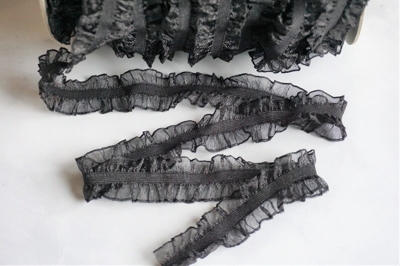 May include: Black ruffled elastic trim, perfect for sewing and crafting projects. The trim features a delicate, sheer fabric with a ruffled edge and a sturdy elastic base. The trim is shown in a roll and in several cut pieces.
