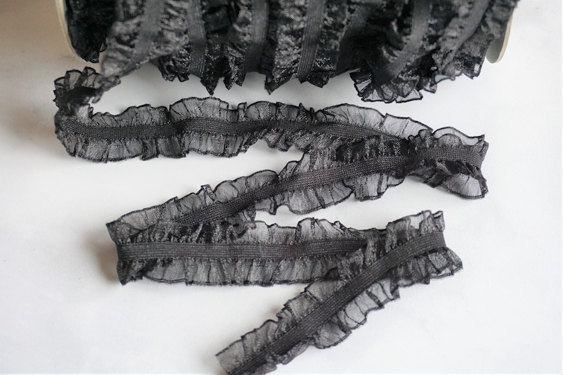 May include: Black ruffled elastic trim, perfect for sewing and crafting projects. The trim features a delicate, sheer ruffle along the edge, adding a touch of elegance. The trim is shown in a roll and in several cut pieces.