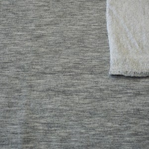 May include: A close-up shot of two fabric swatches. The larger swatch is a heather gray knit fabric. A smaller, off-white, textured fabric is in the upper right corner. The fabrics are likely for apparel or home decor.