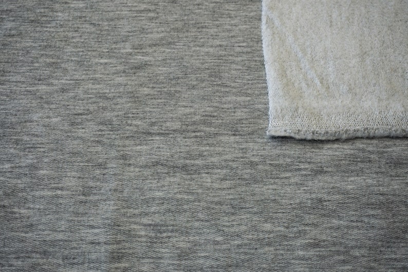 May include: A close-up shot of two fabric swatches. The larger swatch is a heather gray knit fabric. A smaller, off-white, textured fabric is in the upper right corner. The fabrics are likely for apparel or home decor.