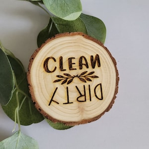 May include: A round wooden sign with the words "CLEAN" and "DIRTY" burned into the wood. A decorative sprig is between the words. The sign is made from a cross-section of a tree branch, with the bark still attached.