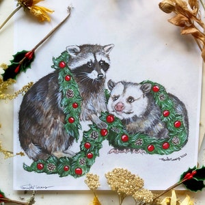 Print of a raccoon stands on his back legs crouching forward, holding a large green garlands of pine, pine cones, and red globe ornaments. To his left is an opossum sitting and smiling with the garland draped over his neck