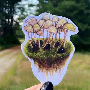 May include: A sticker featuring a watercolor illustration of a cluster of yellow mushrooms with black eyes peeking out from beneath them. The mushrooms are growing on a patch of green moss.