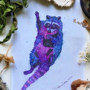 May include: A watercolor painting of a raccoon with a galaxy pattern. The raccoon is depicted with its paws raised in the air, as if it is floating. The painting is signed "Traubacious 2022" in the bottom right corner.
