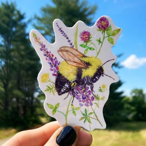 May include: A sticker featuring a bumblebee with black and yellow stripes, perched on purple and white flowers. The sticker is on a white background and has a slightly irregular shape.