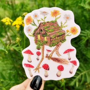 May include: A whimsical sticker featuring a small wooden cabin with a mossy roof perched atop spindly legs. The cabin is surrounded by white and yellow flowers, and the legs are standing on a bed of red and white mushrooms.