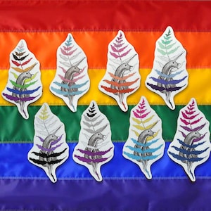 May include: A collection of eight stickers featuring a unicorn-like creature with a fern-shaped body. Each sticker has a different color scheme, with the background showing a rainbow flag.