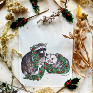 Print of a raccoon stands on his back legs crouching forward, holding a large green garlands of pine, pine cones, and red globe ornaments. To his left is an opossum sitting and smiling with the garland draped over his neck