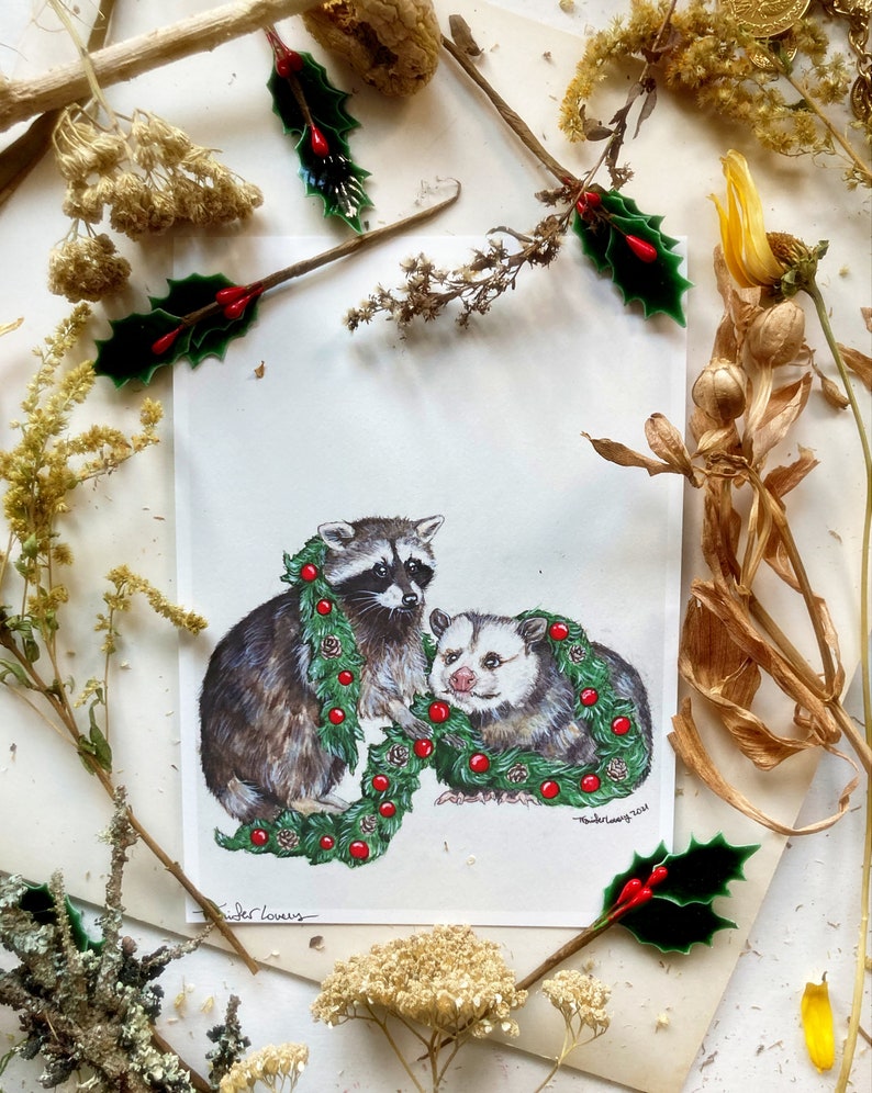 Print of a raccoon stands on his back legs crouching forward, holding a large green garlands of pine, pine cones, and red globe ornaments. To his left is an opossum sitting and smiling with the garland draped over his neck
