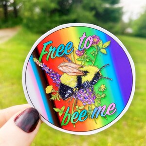 May include: A round sticker with a rainbow background and a bumblebee surrounded by flowers. The text "Free to bee me" is written in a colorful font.