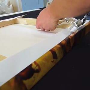 Peut inclure: Une personne utilise un pistolet &agrave; agrafer pour fixer une toile &agrave; un cadre en bois. La toile est imprim&eacute;e d'une image color&eacute;e.