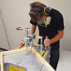 Peut inclure: Une personne portant un t-shirt noir et une protection auditive utilise une machine pour assembler des cadres photo en bois. La machine a une base m&eacute;tallique et une s&eacute;rie de pinces qui maintiennent les pi&egrave;ces du cadre ensemble. Le cadre est en bois clair.