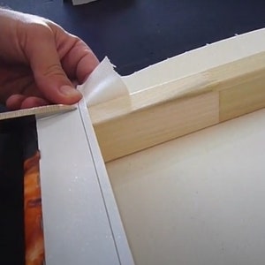 Peut inclure: Une main utilise des ciseaux pour couper l'exc&eacute;dent de toile autour du bord d'un cadre en bois. La toile est blanche et le cadre est brun clair.
