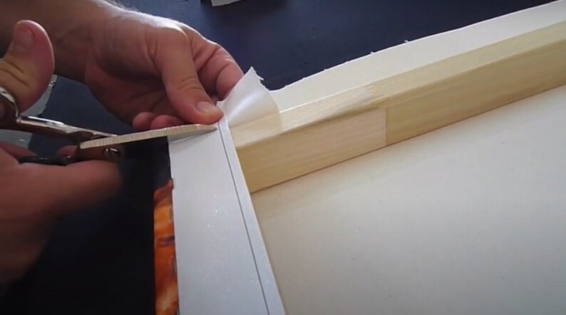 Peut inclure: Une main utilise des ciseaux pour couper l'exc&eacute;dent de toile autour du bord d'un cadre en bois. La toile est blanche et le cadre est brun clair.