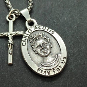 May include: Silver chain necklace with a silver cross pendant and a silver oval pendant with a portrait of Carlo Acutis and the text "CARLO ACUTIS Pray For US".