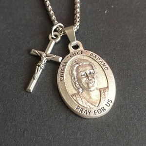 May include: Silver chain necklace with a silver pendant featuring a portrait of Chiara Luce Badano and the text "CHIARA LUCE BADANO PRAY FOR US". A silver crucifix is attached to the chain.