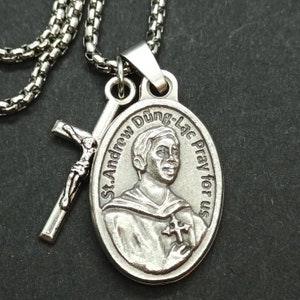Peut inclure: Collier avec pendentif en argent avec un charme ovale en argent représentant le portrait d'un homme avec le texte "St. Andrew Dung-Lac pray for us". Le charme est attaché à une croix en argent.