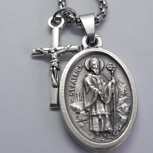 May include: Silver tone necklace with a St. Patrick's medal and a small cross pendant. The medal features a detailed image of St. Patrick holding a staff and a shamrock.