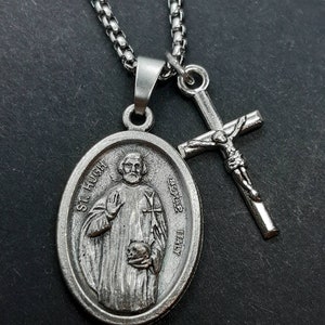 May include: Silver chain necklace with two pendants. One pendant is a round silver medallion with the image of a man and the text "ST. HUGH" and "BUYERS ITALY". The other pendant is a silver cross with a figure of Jesus Christ.