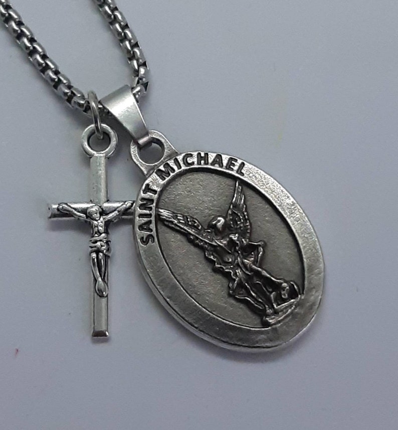K&ouml;nnte beinhalten: Eine silberfarbene Halskette mit zwei Anh&auml;ngern: ein Kruzifix und ein ovales Medaillon mit der Aufschrift "SAINT MICHAEL" und einem Engelbild. Die Kette hat ein quadratisches Glieddesign.