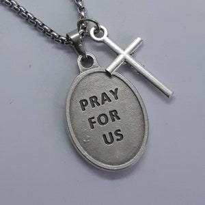 K&ouml;nnte beinhalten: Silberfarbene Halskette mit einem Anh&auml;nger mit der Aufschrift "PRAY FOR US" und einem kleinen Kreuzanh&auml;nger. Der ovale Anh&auml;nger und das Kreuz sind an einer Kette befestigt. Die Halskette wird vor wei&szlig;em Hintergrund gezeigt.