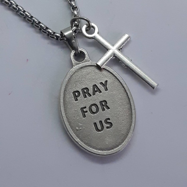 K&ouml;nnte beinhalten: Silberfarbene Halskette mit einem Anh&auml;nger mit der Aufschrift "PRAY FOR US" und einem kleinen Kreuzanh&auml;nger. Der ovale Anh&auml;nger und das Kreuz sind an einer Kette befestigt. Die Halskette wird vor wei&szlig;em Hintergrund gezeigt.