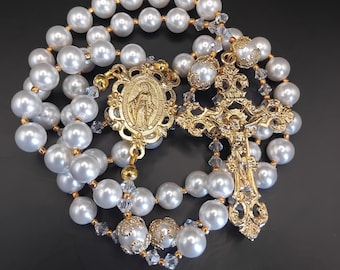 Può includere: Un rosario con perle bianche finte alternate a piccoli cristalli trasparenti sfaccettati. Il pezzo include un crocifisso in metallo dorato con dettagli in filigrana e una medaglia ovale centrale che raffigura una figura in rilievo.