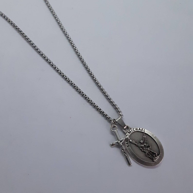 K&ouml;nnte beinhalten: Silberfarbene Halskette mit einem Anh&auml;nger mit einem ovalen Saint Michael Medaillon und einem Schwert-Charm. Die Kette hat ein Box-Link-Design. Der Anh&auml;nger zeigt Saint Michael mit dem Text "SAINT MICHAEL".