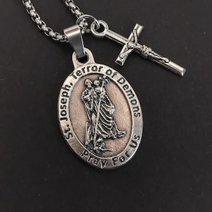 Pode incluir: Colar prateado com um pingente oval e uma cruz. O pingente apresenta uma imagem de duas figuras e o texto "St. Joseph, Terror of Demons, Pray For Us". A cruz tem um design pequeno e detalhado.
