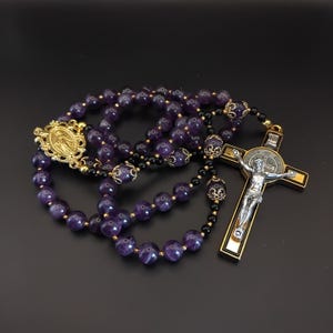May include: A rosary with purple amethyst beads, black beads, and gold-colored accents. The rosary features a gold-colored crucifix with a silver figure of Jesus and a gold-colored medallion.
