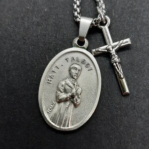 Peut inclure: Collier en chaîne argentée avec un pendentif ovale en argent représentant l'image d'un homme et le texte "MATT. TALBOT ITALY". Une croix en argent avec une figurine de Jésus-Christ est attachée à la chaîne.