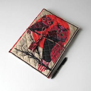 May include: A beige fabric journal cover with a quilted design features two owls with red, black and pink patterns against a red sun. The owls are perched on a branch with black leaves.