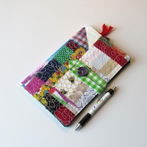 May include: A patchwork fabric case in a variety of colours and patterns, with a button fastening and a red tassel. The case is next to a black and silver pen. The case is made of various fabrics, including floral and plaid.