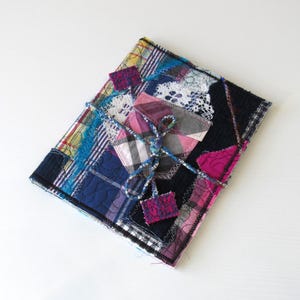 May include: A patchwork fabric journal with a blue, pink and white colour scheme. The journal is tied closed with a blue and white cord and has a pink fabric flower accent.
