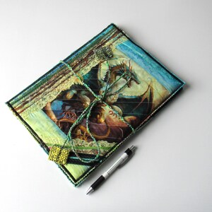 May include: A quilted book cover with a dragon illustration. The cover combines green, blue, and brown fabrics, with a decorative lace trim. A green cord wraps around the cover, and a pen is placed beside it.