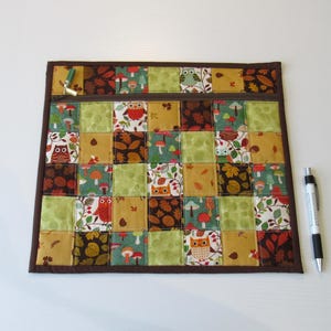 May include: A square quilted pouch featuring a patchwork design of autumn-themed fabrics in shades of brown, green, orange, and cream. The pattern includes owls, mushrooms, and leaves. A dark brown zip runs across the top, and a pen is placed beside it.