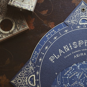 May include: A detailed image of a vintage planisphere and a decorative box. The planisphere is navy blue with gold accents and astronomical details. The box is wooden with intricate designs. The items are on a dark, patterned surface.