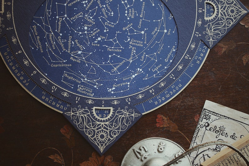 May include: A detailed celestial chart with constellations, zodiac signs, and star names, printed on dark blue paper. The chart features intricate designs and a circular scale, with the text "FEBRUARY" and "NOVEMBER" visible. The chart is set on a dark wooden surface.