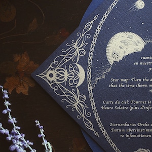 May include: A navy blue star map with gold foil celestial illustrations, including a moon, sun, and stars. The map has text in multiple languages and is a unique piece of art.