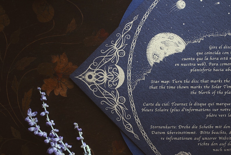 May include: A navy blue star map with gold foil celestial illustrations, including a moon, sun, and stars. The map has text in multiple languages and is a unique piece of art.
