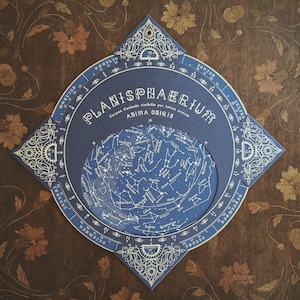 May include: A blue and gold Planisphaerium, a star chart, with the text "PLANISPHAERIUM" and "ANIMA OUIIS". The chart features a detailed depiction of constellations against a dark blue background. The chart is set on a dark brown wooden surface with floral designs.