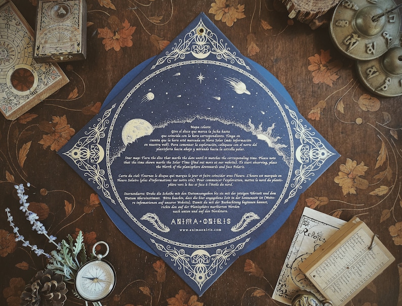 May include: A dark blue, star-themed card with ornate gold detailing, featuring celestial illustrations of the moon, sun, and stars. The card includes text in multiple languages and is surrounded by other vintage-style items, including a compass and a small wooden box.
